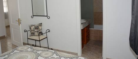 A bedroom with a bed, a chair, a mirror, and a bathroom visible through an open door.