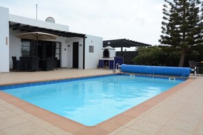 A swimming pool with a patio area, a covered seating area, and a large tree in the background. - Villa julima 110 m2 3 ch AC everywhere wifi broadband, tv uk heated pool (Playa Blanca)