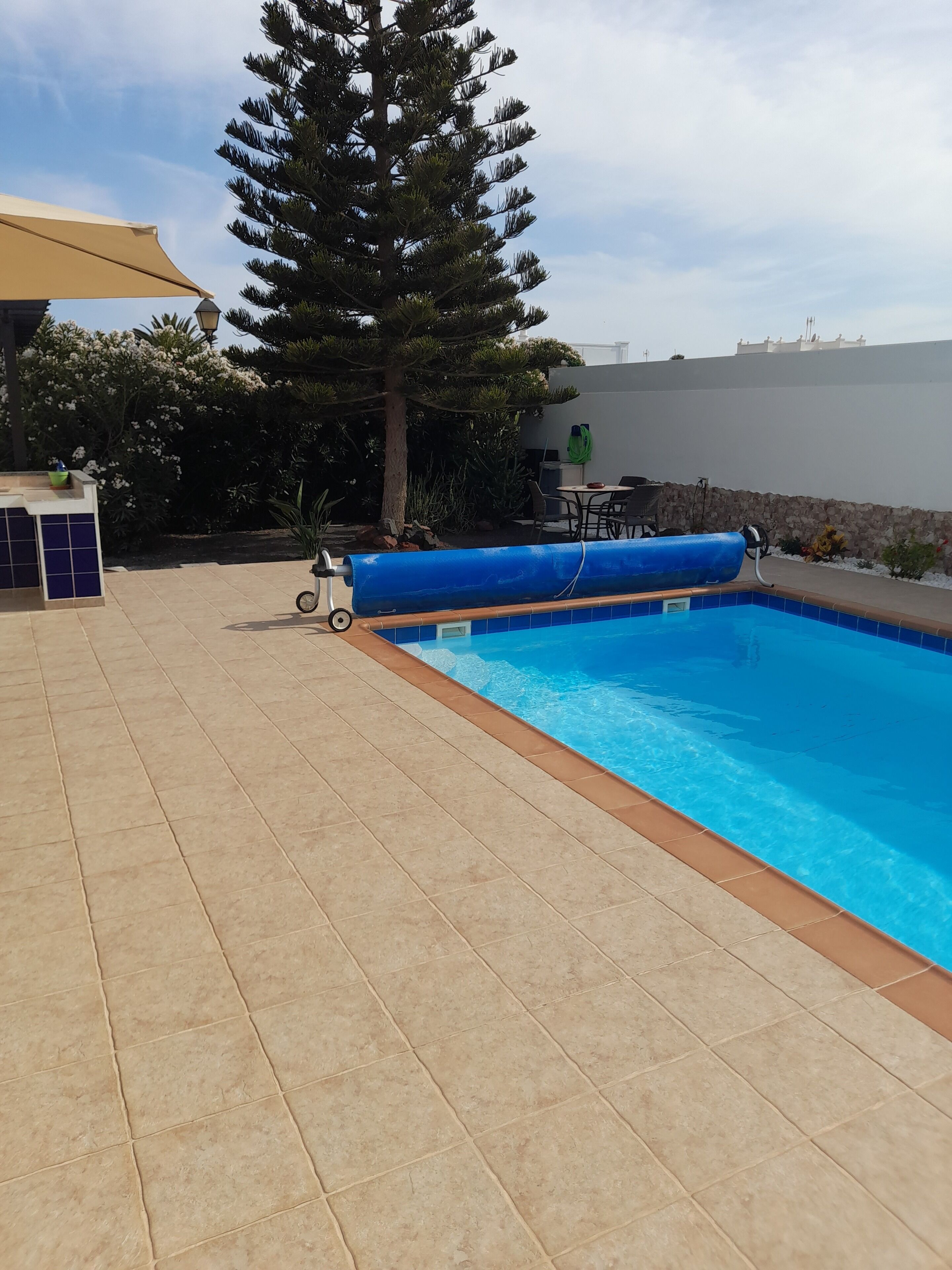Une piscine avec une terrasse carrelée, un espace détente avec une table et des chaises, et un grand arbre en arrière-plan.