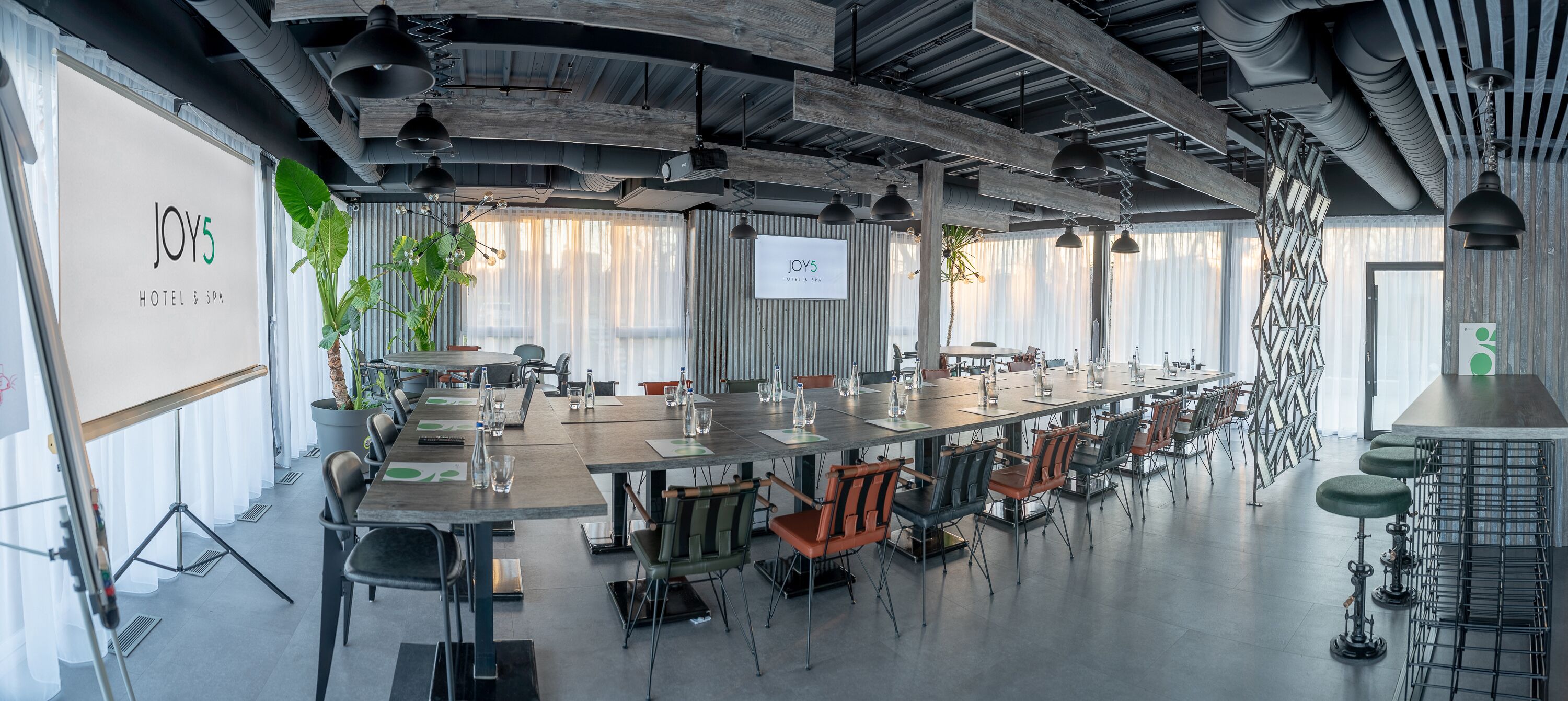 A spacious conference room with long tables and chairs, a large screen, and a potted plant.