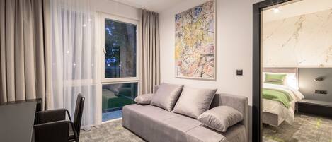 A modern hotel room with a grey sofa, a dining table, and a large window with curtains.