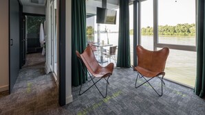 Une chambre d’hôtel moderne dotée d’une grande fenêtre, de deux fauteuils en cuir, d’un téléviseur à écran plat et offrant une vue sur une étendue d’eau.
