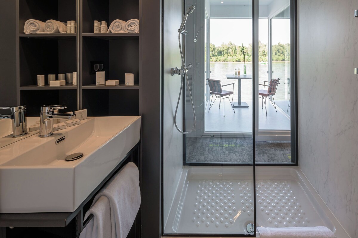 Un baño moderno con ducha envuelta en vidrio, tocador con lavamanos y repisas con toallas y artículos de aseo.