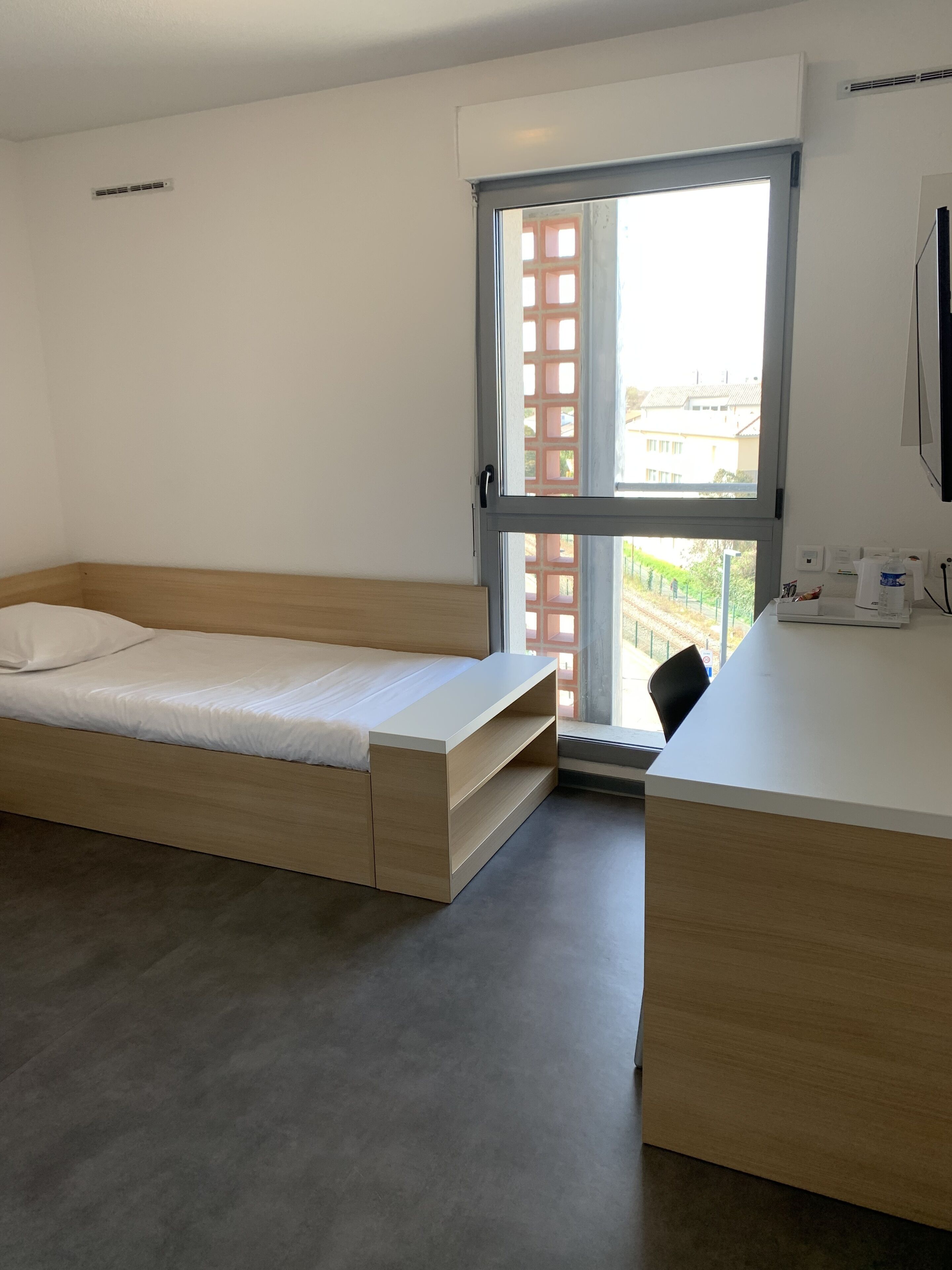 Una habitación con cama, escritorio y ventana con vistas a edificios y vegetación.