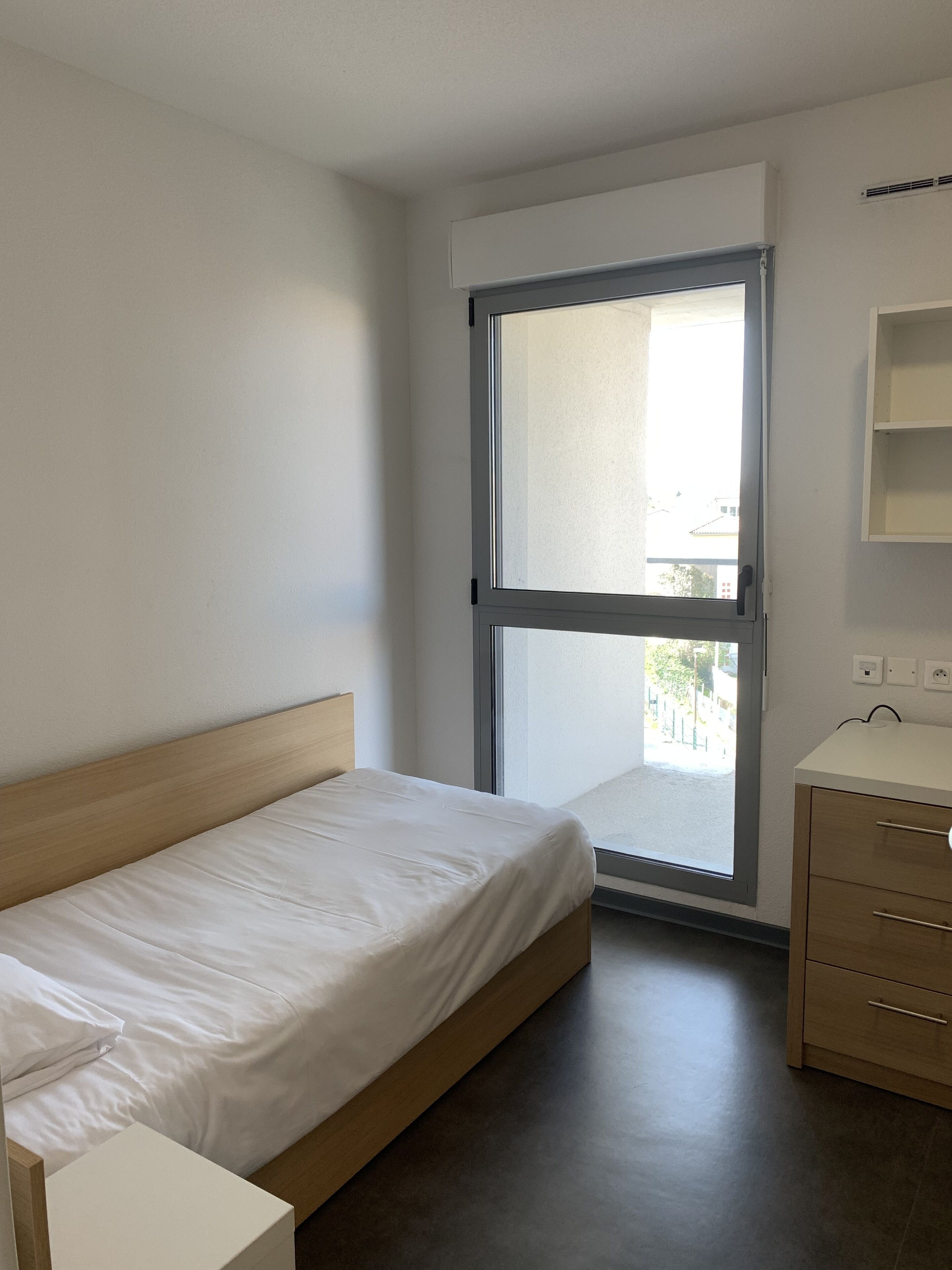 Ein kleines, sauberes Zimmer mit einem Einzelbett, einem Fenster, einem Nachttisch und einer kleinen Kommode.