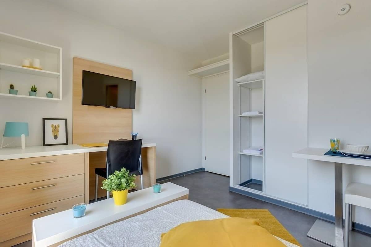 A modern, minimalistic bedroom with a bed, a desk, a chair, a TV mounted on the wall, and built-in shelving.