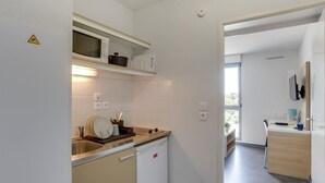 A compact kitchen with a microwave, sink, and a small refrigerator. There is a shelf above the microwave and a countertop with a dish rack. The kitchen leads to a dining area with a table and chairs. - Apparteo Toulouse (Toulouse)