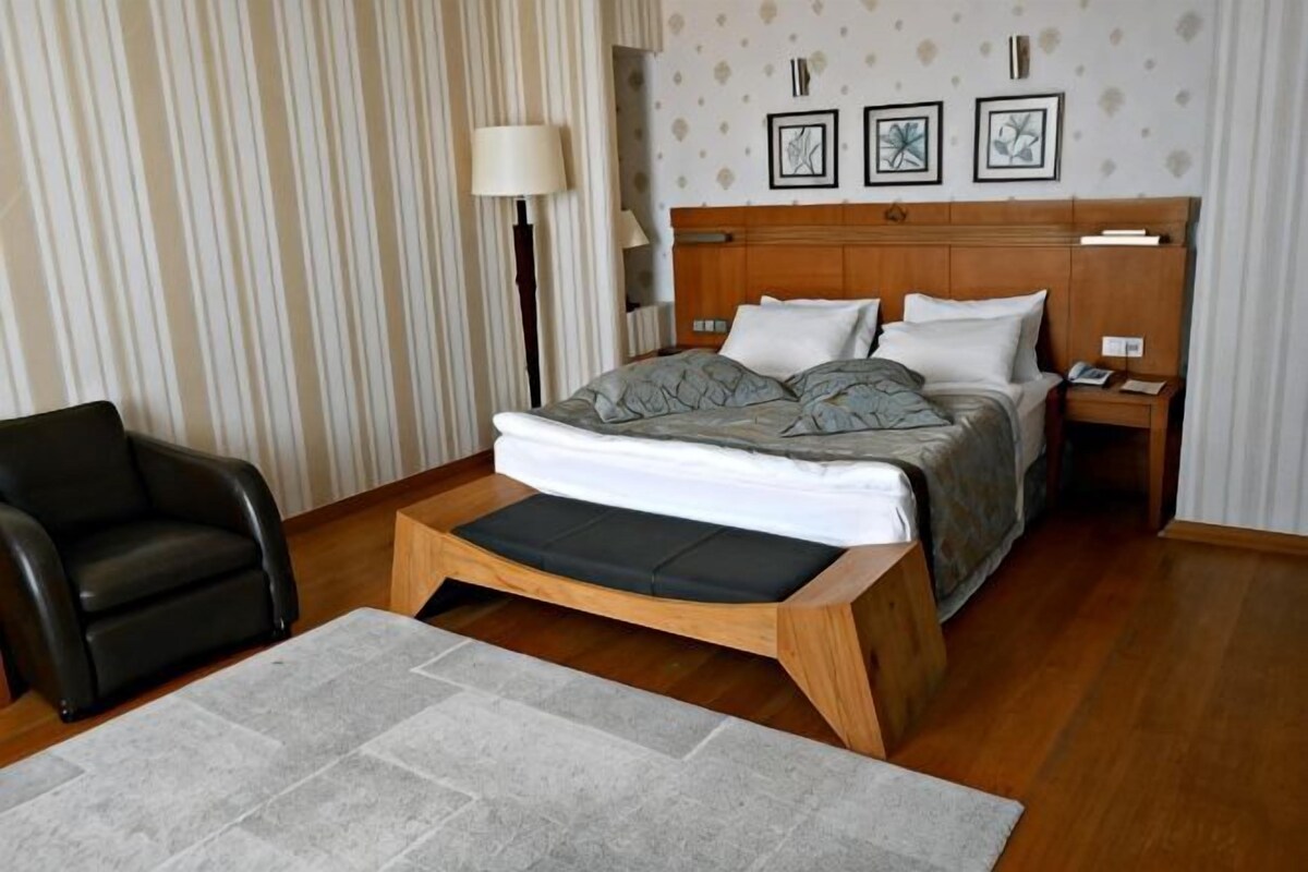 A bedroom with a bed, a chair, a bedside table, a lamp, and a wall with framed pictures.