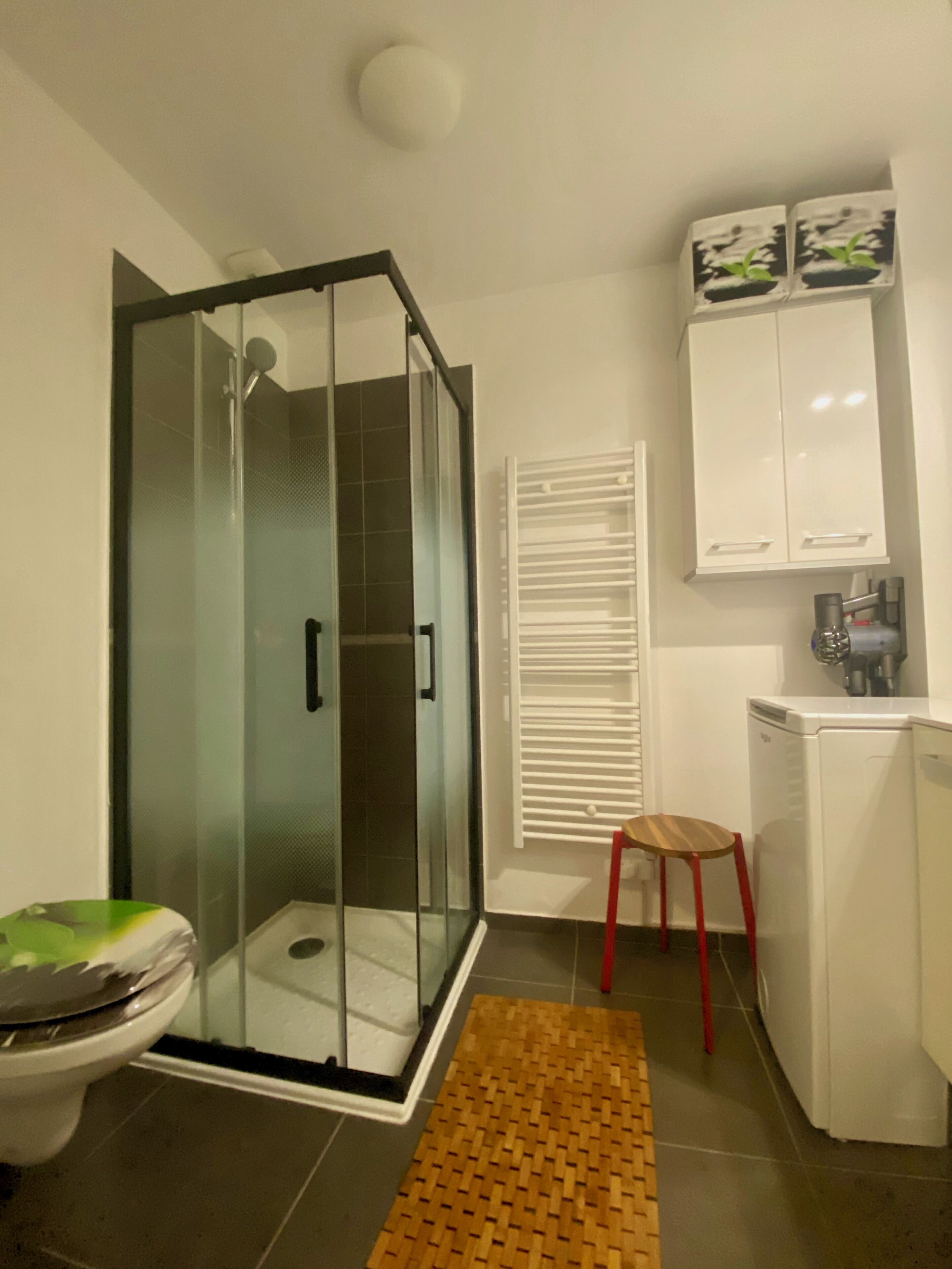 A bathroom with a glass-enclosed shower, a toilet, a wooden stool, a white cabinet, and a radiator.