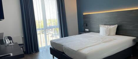 Una habitación de hotel moderna con una cama grande, un escritorio y vistas al exterior a través de un amplio ventanal con cortinas.