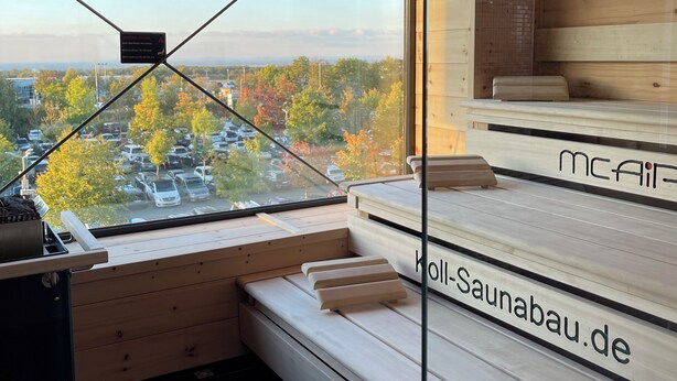 Ein modernes Interieur mit einem großen Fenster, das einen Blick auf einen Parkplatz und Bäume bietet. Holzbanken und ein kleiner Tisch mit Wasserkocher sind vorhanden.