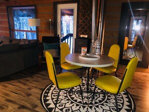 Quarto aconchegante com mesa redonda, quatro cadeiras amarelas, tapete estampado e janela com vista para árvores.