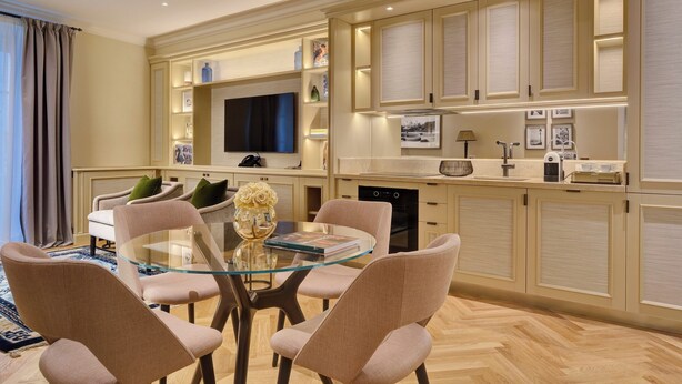 Een moderne eethoek met een glazen tafel, beklede stoelen, een televisie en keukenkastjes met inbouwverlichting.