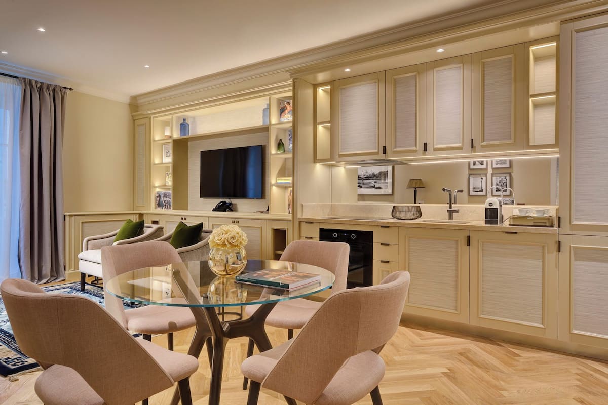 Un comedor moderno con una mesa de vidrio y sillas acolchadas, un televisor y armarios de cocina con iluminación empotrada.