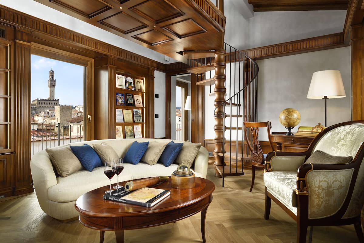 Amplia sala de estar con escalera de madera, sofá con cojines azules, mesa de centro de madera con un libro y copas de vino, y vista panorámica de la ciudad a través de grandes ventanales.