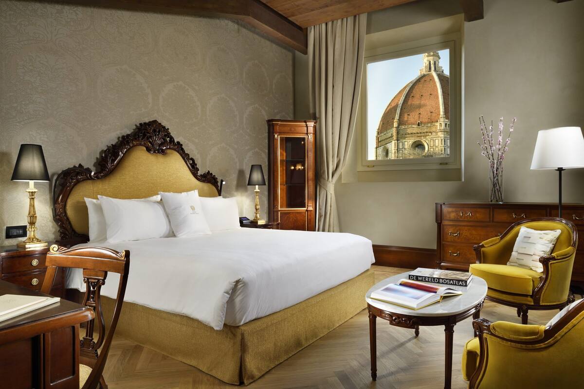 Habitación de hotel con una cama grande, muebles de madera, un escritorio, una silla, una lámpara y vista a un edificio a través de una ventana.