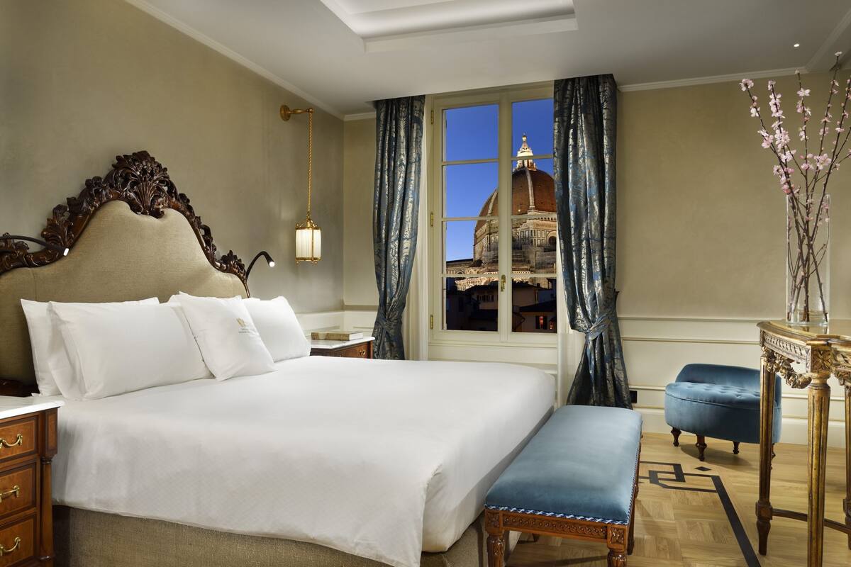 Un dormitorio amplio con una cama grande, una mesita de noche de madera, un banco y vistas a un edificio histórico por la ventana.
