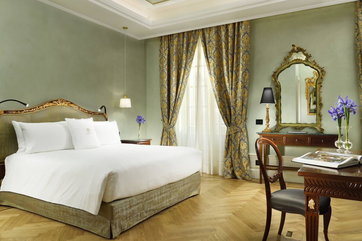 Un dormitorio elegante con una cama grande, un escritorio de madera con una silla, un espejo y un jarrón con flores.
