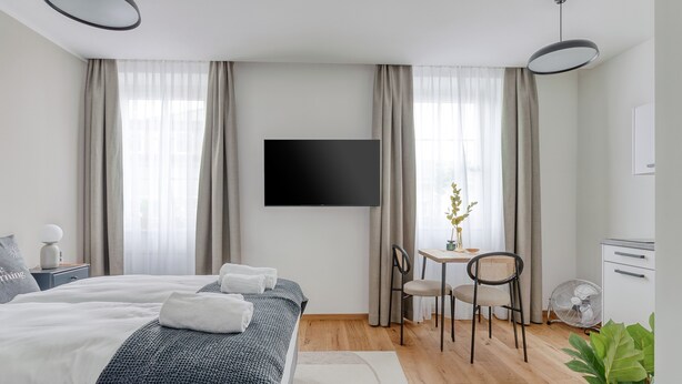 Quarto de hotel moderno com uma cama grande, uma escrivaninha com duas cadeiras, uma televisão de tela plana e uma pequena mesa com uma planta.