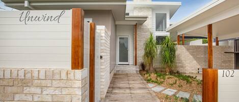 Una casa moderna con una terraza de madera que da a una puerta, un muro de ladrillo y un letrero con el número 102.