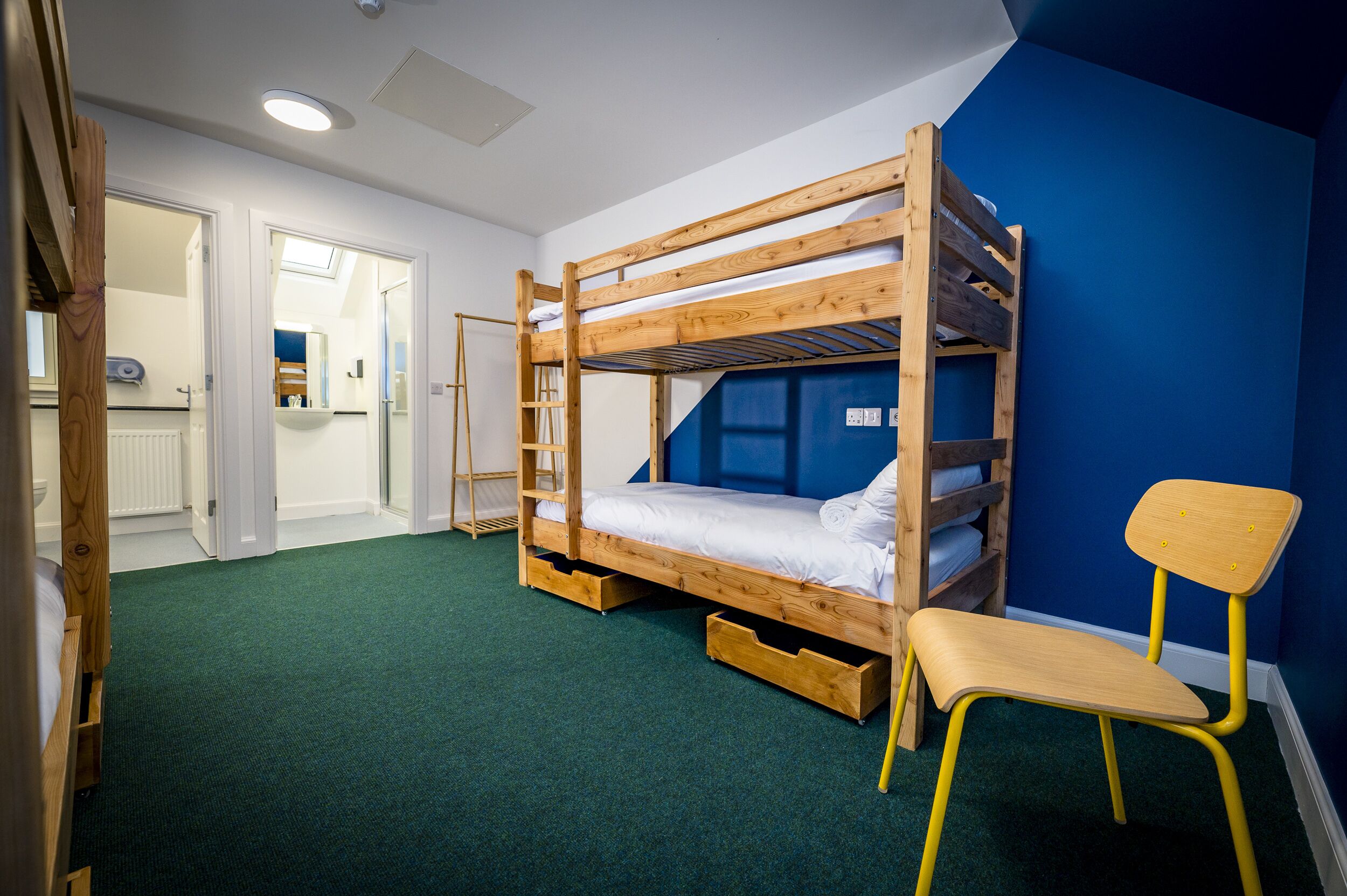 Un letto a castello con cassetti portaoggetti, una sedia di legno e un bagno visibile attraverso una porta aperta.