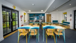 A modern kitchen with blue cabinets and a central island, a dining area with blue and yellow chairs, and a large window with a view of greenery.