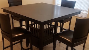 Une table de salle à manger rectangulaire, entourée de quatre chaises, chacune dotée d’un haut dossier, est placée dans un coin d’une pièce dont le sol est carrelé et qui possède une fenêtre avec des stores.
