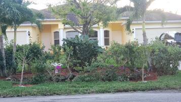 A residential house with a yellow facade, large windows, and a well-maintained garden with various plants and a birdbath.