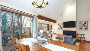 A spacious living room with a wooden dining table, chairs, a fireplace, and a large window overlooking a deck.