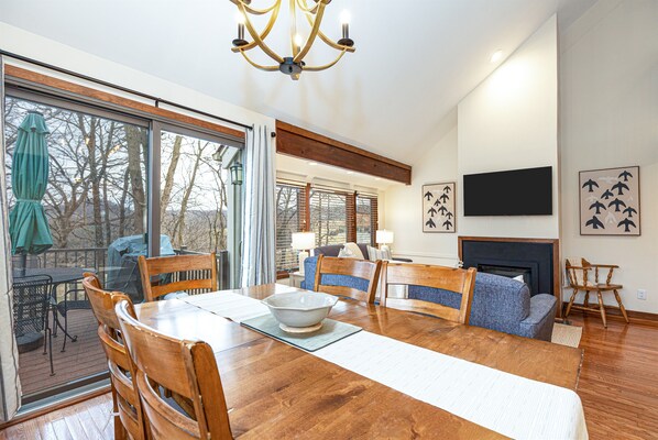 A spacious living room with a wooden dining table, chairs, a fireplace, and a large window overlooking a deck.