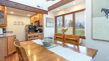 A dining area with a wooden table, chairs, and a bowl. There is a kitchen in the background with wooden cabinets and a refrigerator.