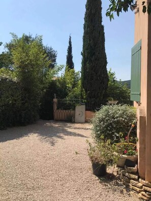 Une allée de gravier menant à une entrée fermée par un portail, entourée d’une végétation luxuriante et de grands arbres.
