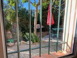 A view through a window with a metal grid, a red cloth hanging, and a garden with a table and chairs.