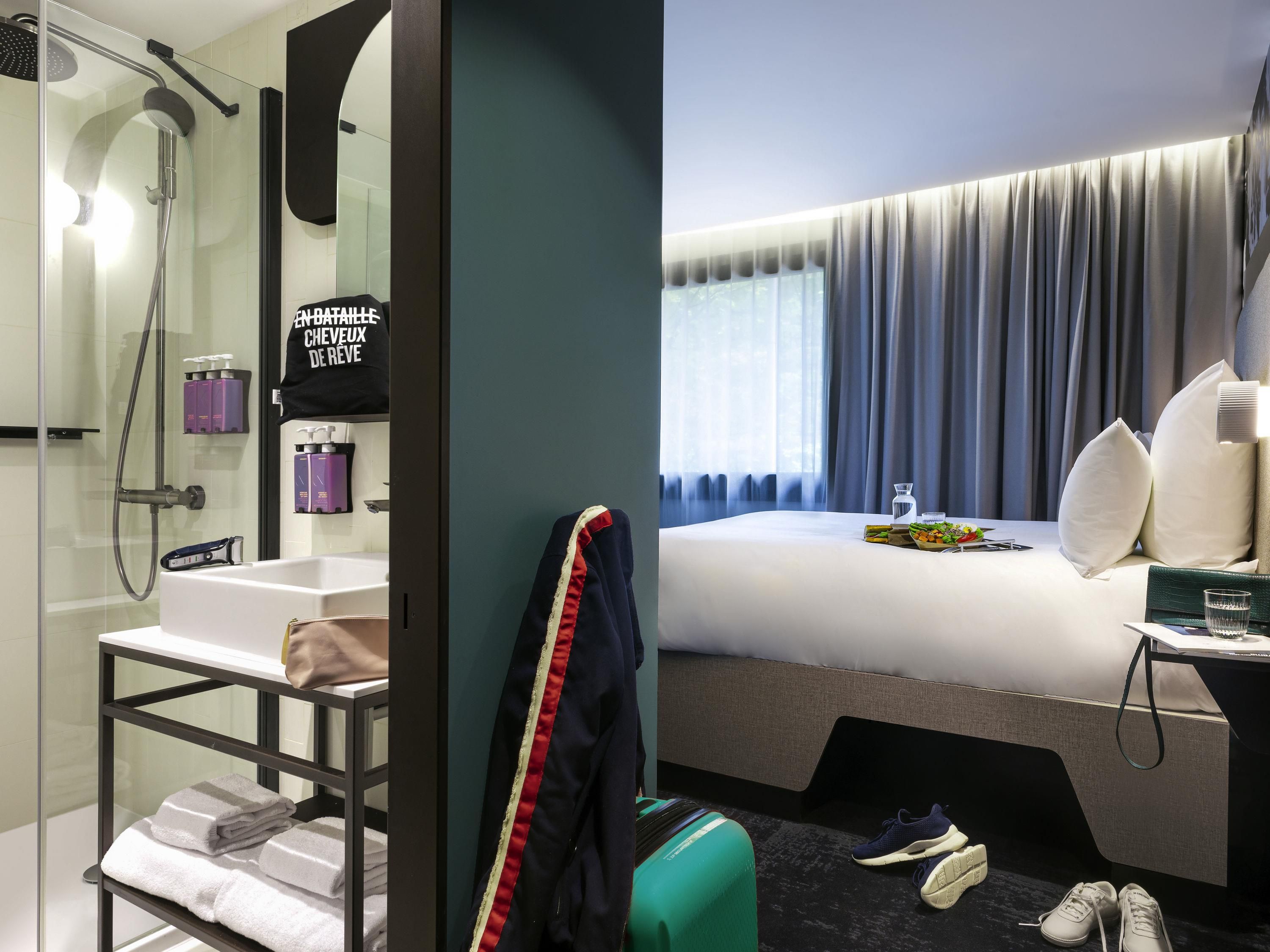 Hotelkamer met douche, bed, nachtkastje, koffer en raam met gordijnen.