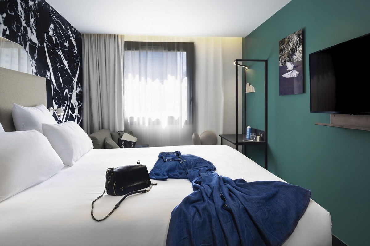 A hotel room with a bed, a flat-screen TV, a desk, and a window with curtains.
