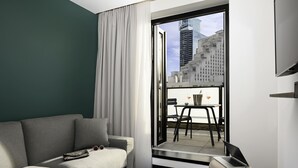 A modern hotel room with a grey sofa, a small table with two glasses, a laptop, and a view of city buildings through a large window. - Tribe Paris La Defense Esplanade (Courbevoie)