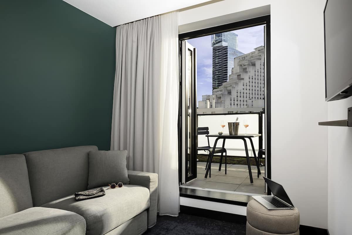 A modern hotel room with a grey sofa, a small table with two glasses, a laptop, and a view of city buildings through a large window.