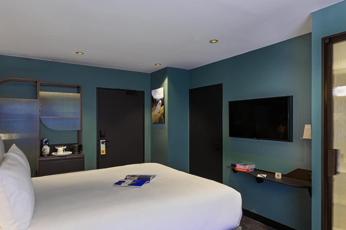 A hotel room with a bed, a TV, a door, and a small shelf with items.