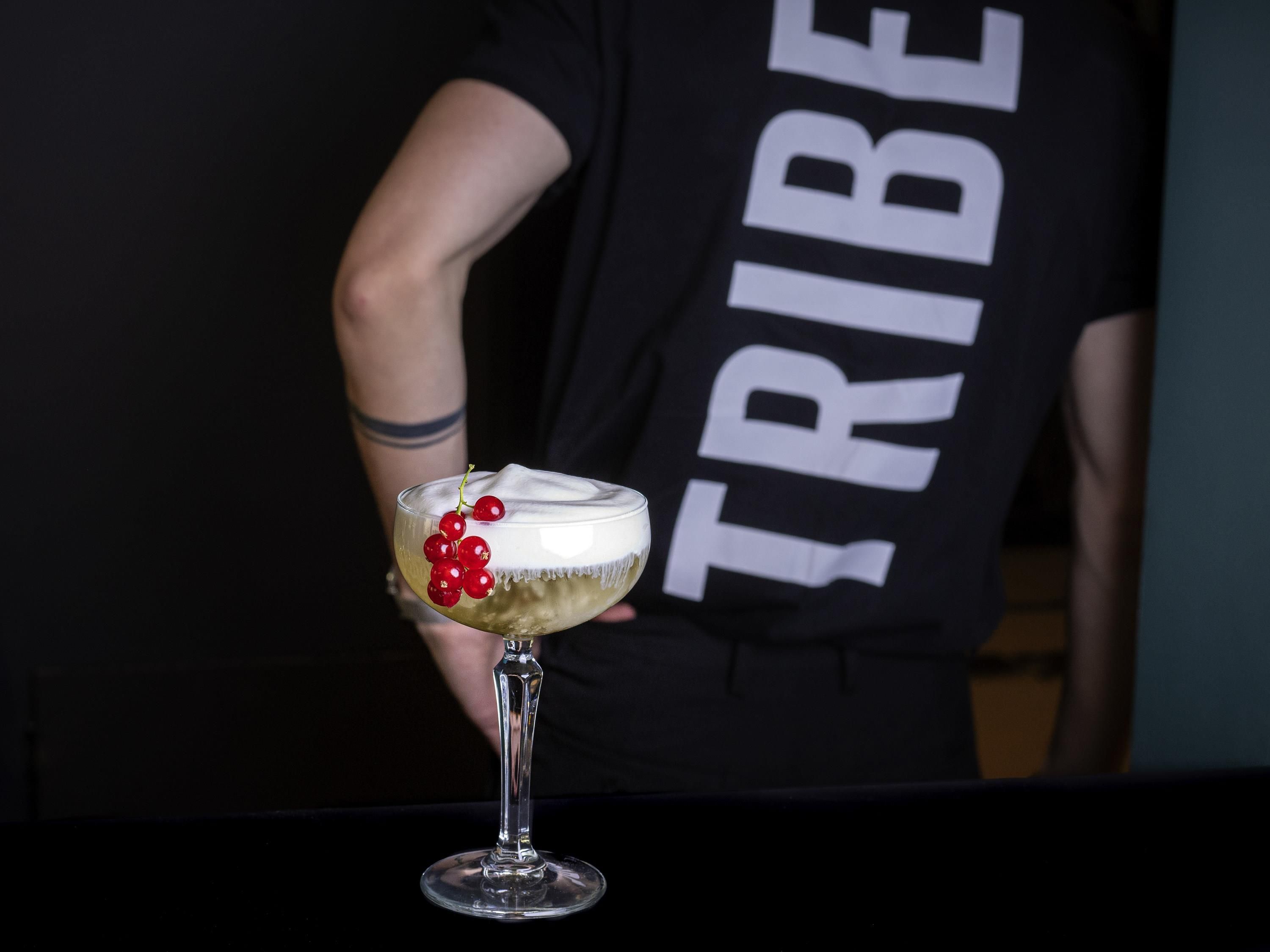 A person holding a cocktail with a garnish, wearing a black shirt with 'TRIBE' printed on it.