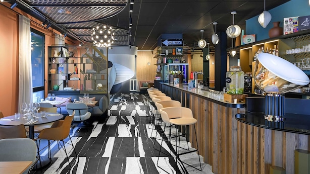 A modern bar area with a checkered floor, bar stools, and a variety of bottles and glasses on the counter. - Tribe Paris La Defense Esplanade (Courbevoie)