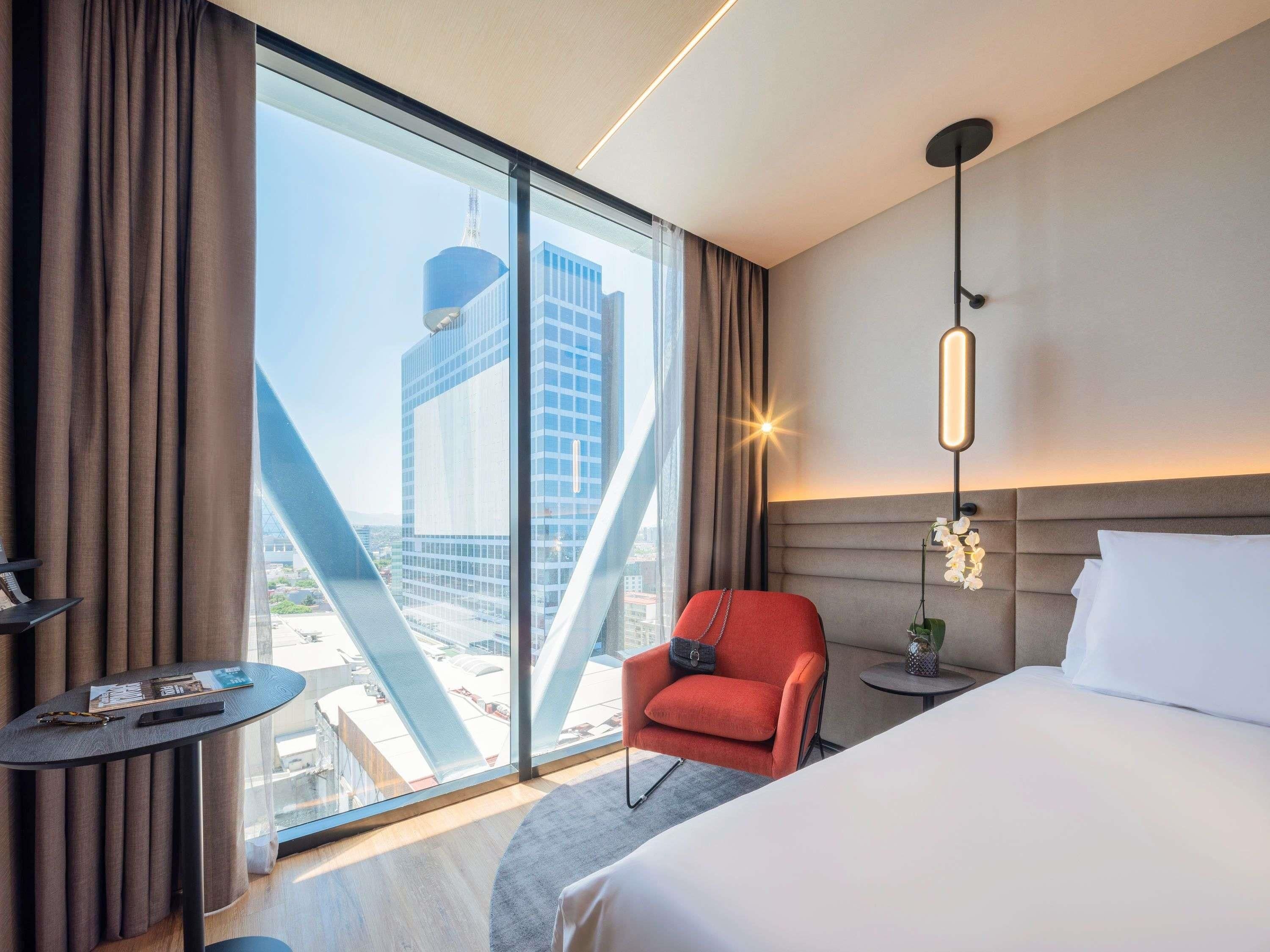 Ein Hotelzimmer mit großem Fenster, einem Bett, einem roten Sessel, einem kleinen Tisch und einem Nachttisch.