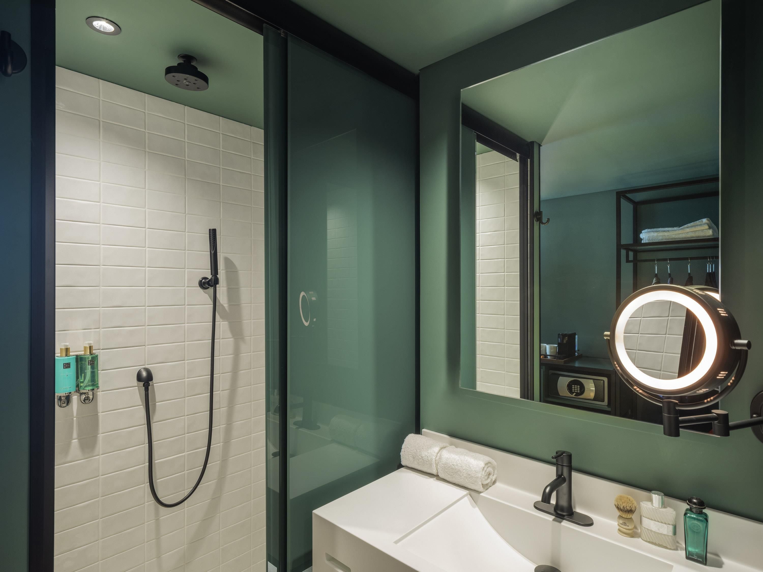 A modern bathroom with a glass-enclosed shower, a white sink, a mirror with a ring light, and toiletries on the counter.