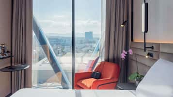 A hotel room with a large window, a bed, a red chair, a small table, and a wall-mounted lamp.