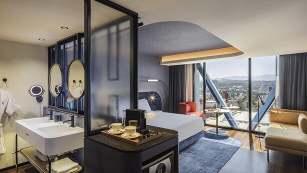 Habitación de hotel moderna con una cama grande, un área de descanso, un escritorio con cafetera, un espejo y vistas a la ciudad.