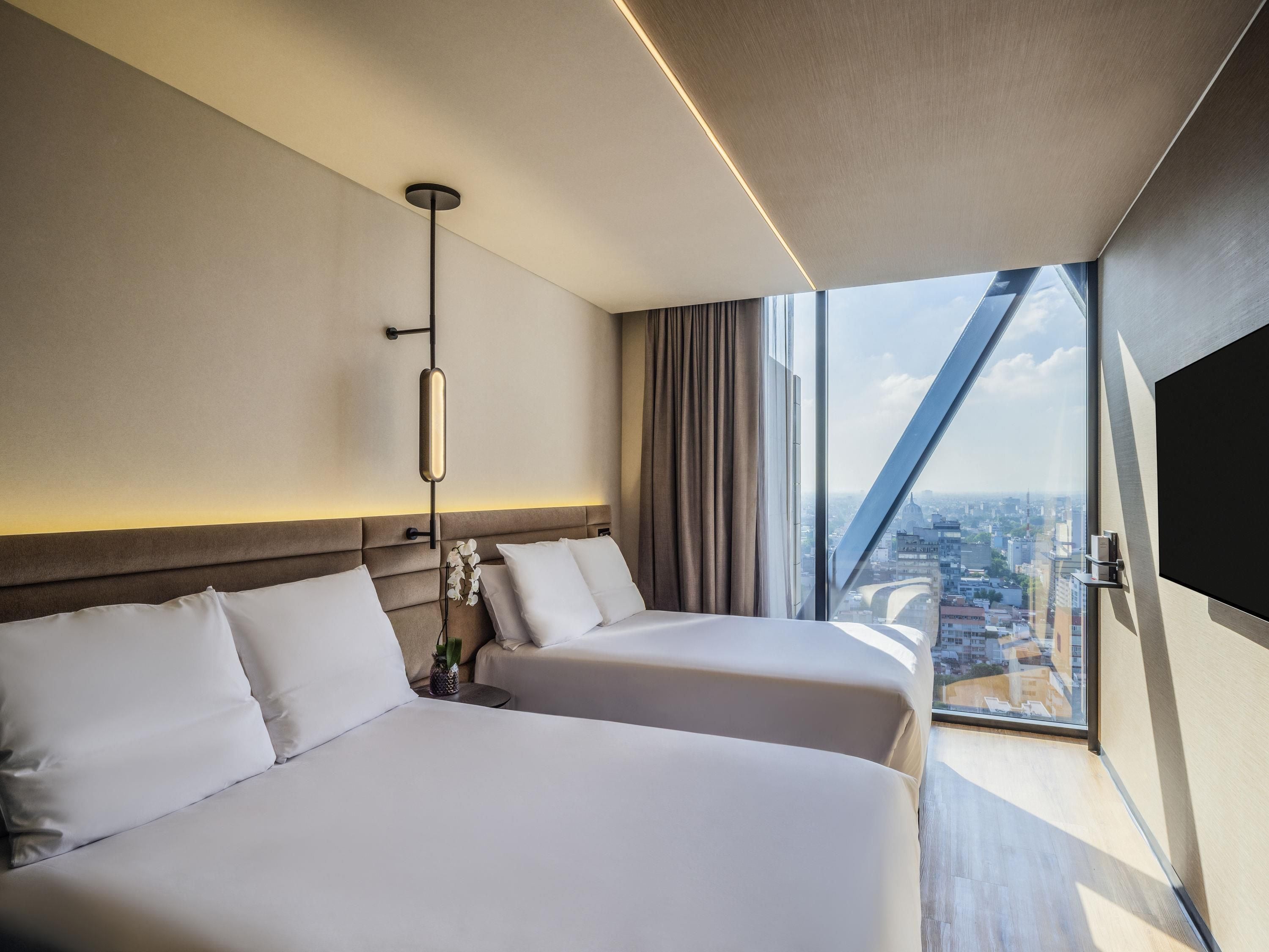 A hotel room with two beds, a large window with a city view, a modern light fixture, and a flat-screen TV mounted on the wall.