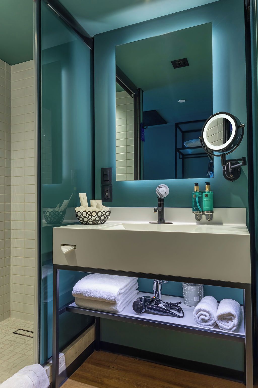 A modern bathroom with a glass partition, a white sink, a mirror with a round light, and a shelf with towels and toiletries.
