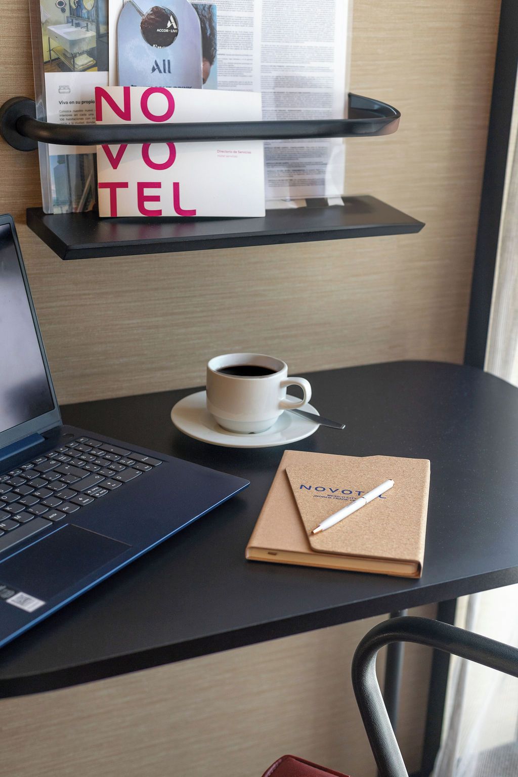 Una laptop sobre una mesa con una taza de café, un cuaderno y un bolígrafo. Hay una estantería con libros y un cartel que dice 'NO VOTE TEL'.