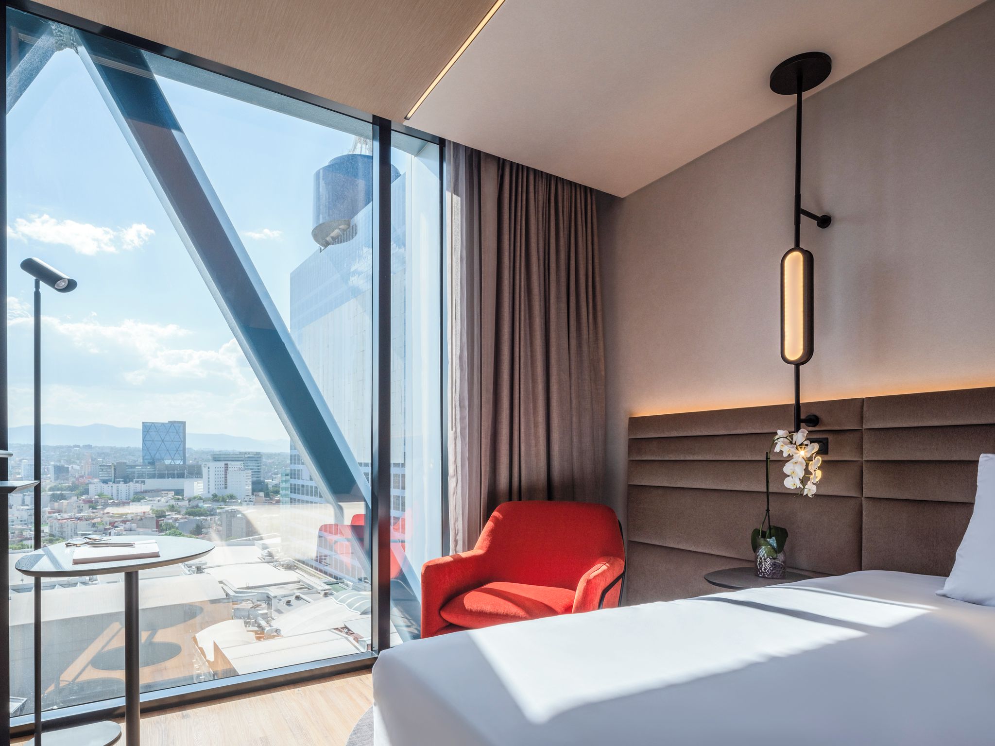 A hotel room with a large window, a red chair, a bed, and a city view.