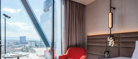A hotel room with a large window, a red chair, a bed, and a city view.