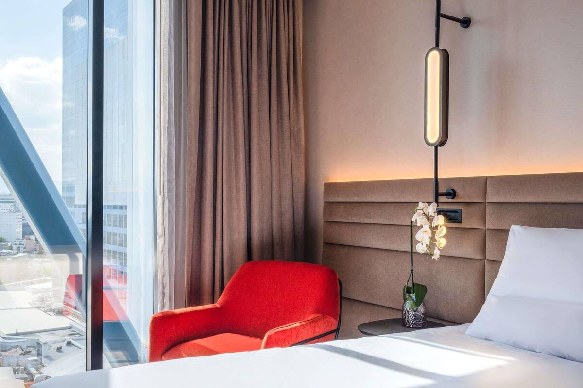 Una habitación de hotel moderna con una cama grande, un sillón rojo, una mesita de noche y una lámpara de pared.
